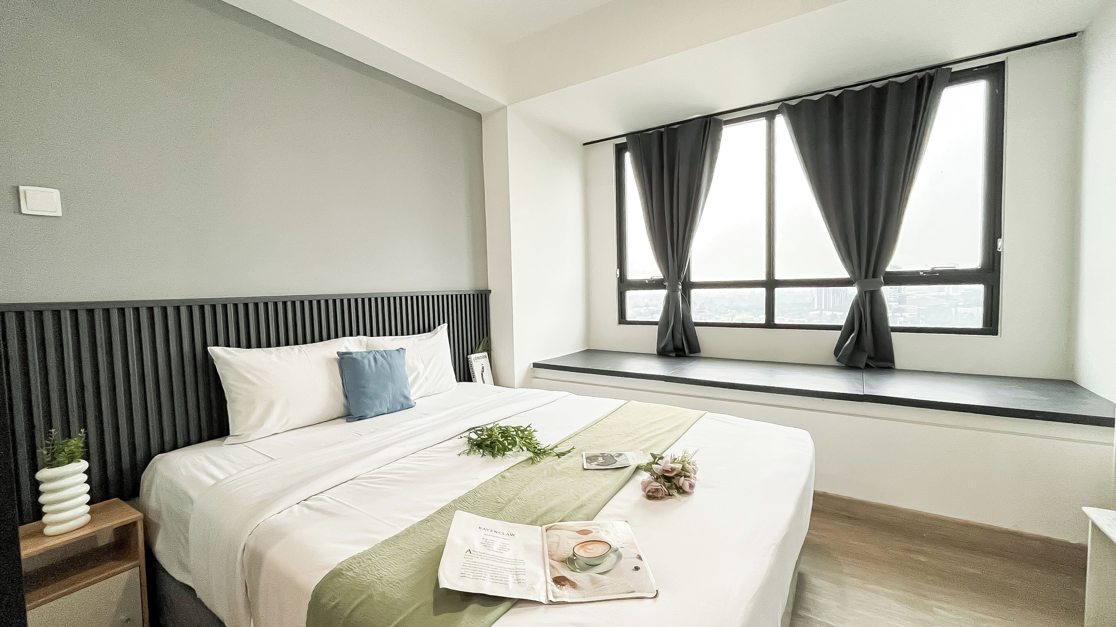A hotel room with a bed, a window with curtains, a bedside table with a plant, and a view of the city.