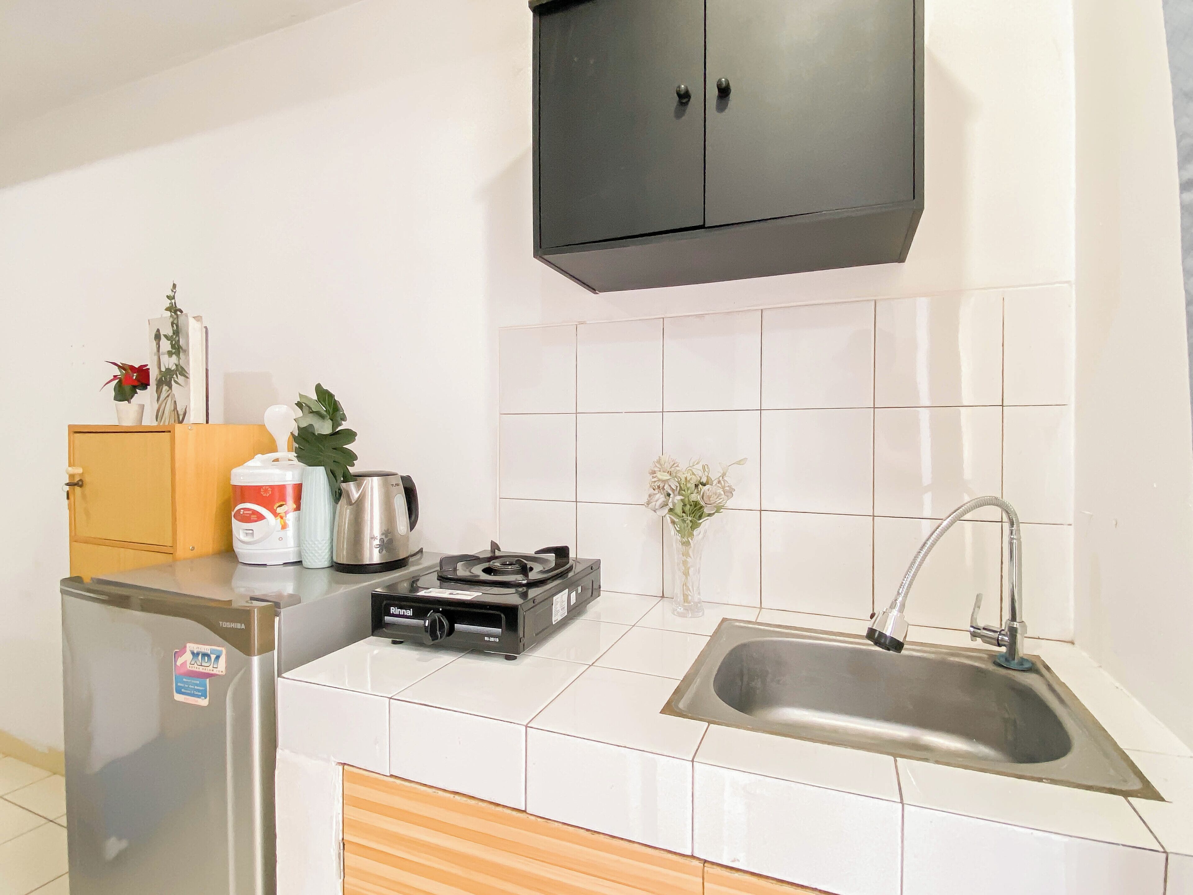 A compact kitchen with white tiles, a stainless steel sink, a black cabinet, a refrigerator, a kettle, and a small potted plant.