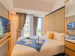 A hotel room with a large bed, a television on a stand, a window with curtains, and a view of buildings outside. - Classic Studio At Tokyo Riverside Pik 2 Apartement (Teluknaga)