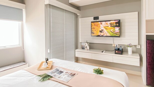 A modern hotel room with a large bed, a mounted TV, an air conditioning unit, and a window with blinds. - Standard Studio Apartment At Lrt City Sentul - Royal Sentul Park (Bogor)
