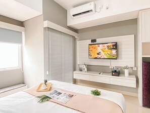 A modern hotel room with a large bed, a mounted TV, an air conditioning unit, and a window with blinds. - Standard Studio Apartment At Lrt City Sentul - Royal Sentul Park (Bogor)