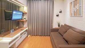 A modern living room with a flat-screen TV, a wooden coffee table, a brown sofa, and a grey curtain. - Modern And Comfy 2Br Apartment Mekarwangi Square Cibaduyut (Bandung)