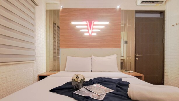 A hotel room with a large bed, a wooden headboard with a V-shaped light fixture, a nightstand with a vase of flowers, and a door to the right. - Modern And Comfy 2Br Apartment Mekarwangi Square Cibaduyut (Bandung)