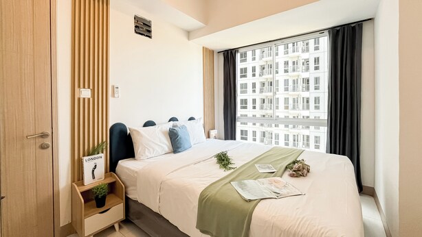 Een hotelkamer met een bed, een nachtkastje met een plant erop, een raam met gordijnen en uitzicht op gebouwen.