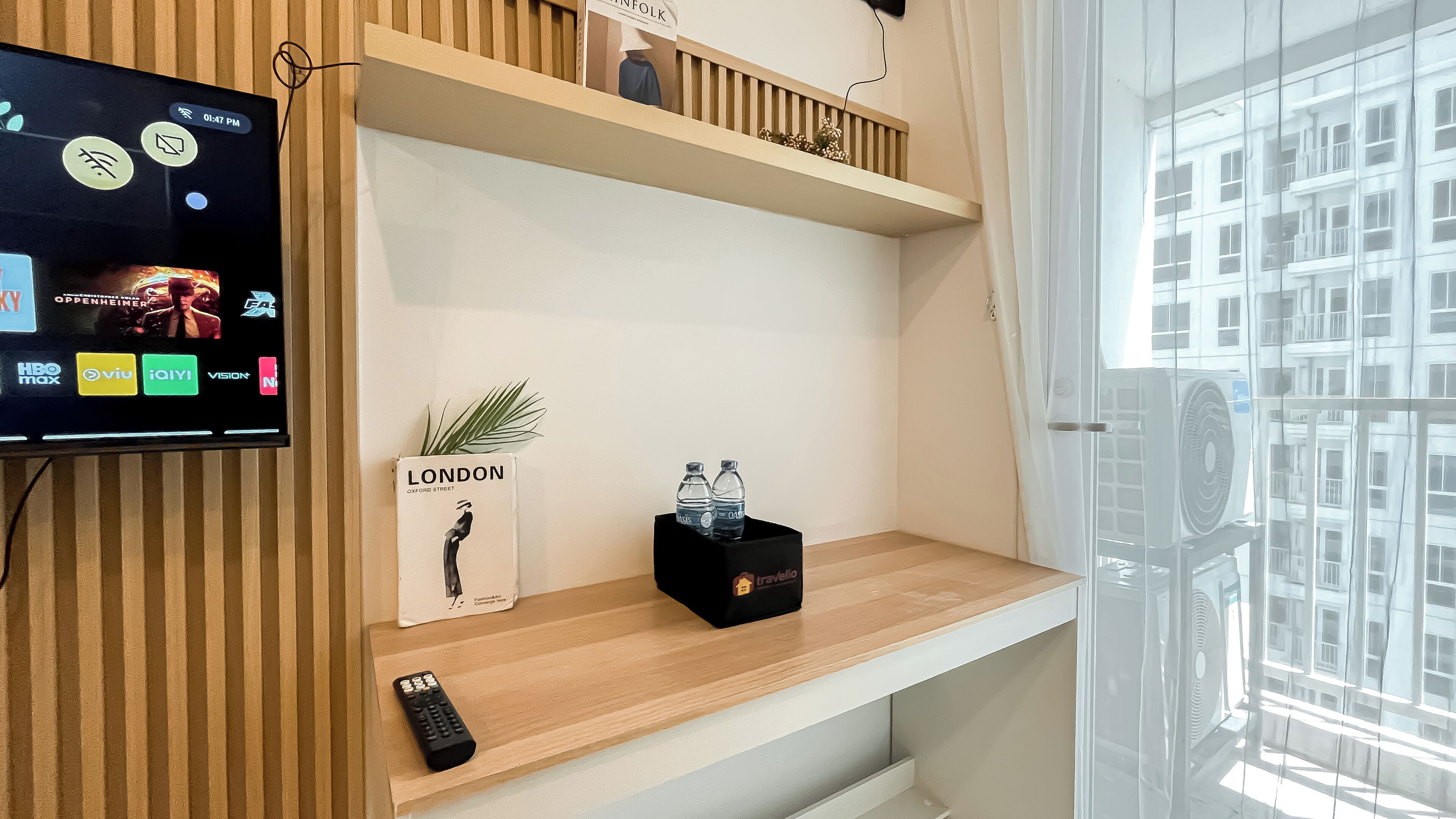 A hotel room with a TV mounted on the wall displaying various channels, a wooden shelf with a plant, a book titled 'LONDON', and a black box with water bottles.