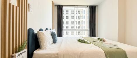 A hotel room with a bed, a window with curtains, a bedside table, and a plant.