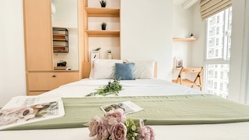 A neatly arranged bedroom with a bed, a wooden wardrobe, a small desk, and a window with a view of buildings.