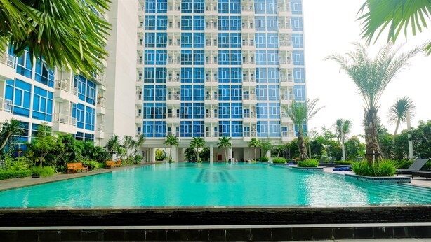 A swimming pool with a view of a high-rise building, surrounded by palm trees and greenery. - Strategic And Spacious 2Br At Capitol Park Residence Apartment (Jakarta)
