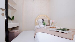 A neatly made bed with a beige bedspread, a small bedside table with a vase of flowers, and a wall-mounted shelf with a plant. - Strategic 2Br Apartment At Green Pramuka City (Jakarta)