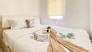 A neatly made bed with a beige blanket, pillows, and a small bedside table with a book and a bottle. - 1Br At Vittoria Residence Near Daan Mogot Mall (Jakarta)