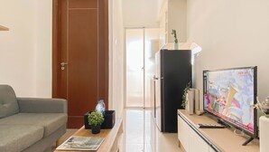 A living room with a sofa, a coffee table with a magazine, a TV on a stand, and a refrigerator. - Trendy 1Br Apartment At Vittoria Residence (Jakarta)