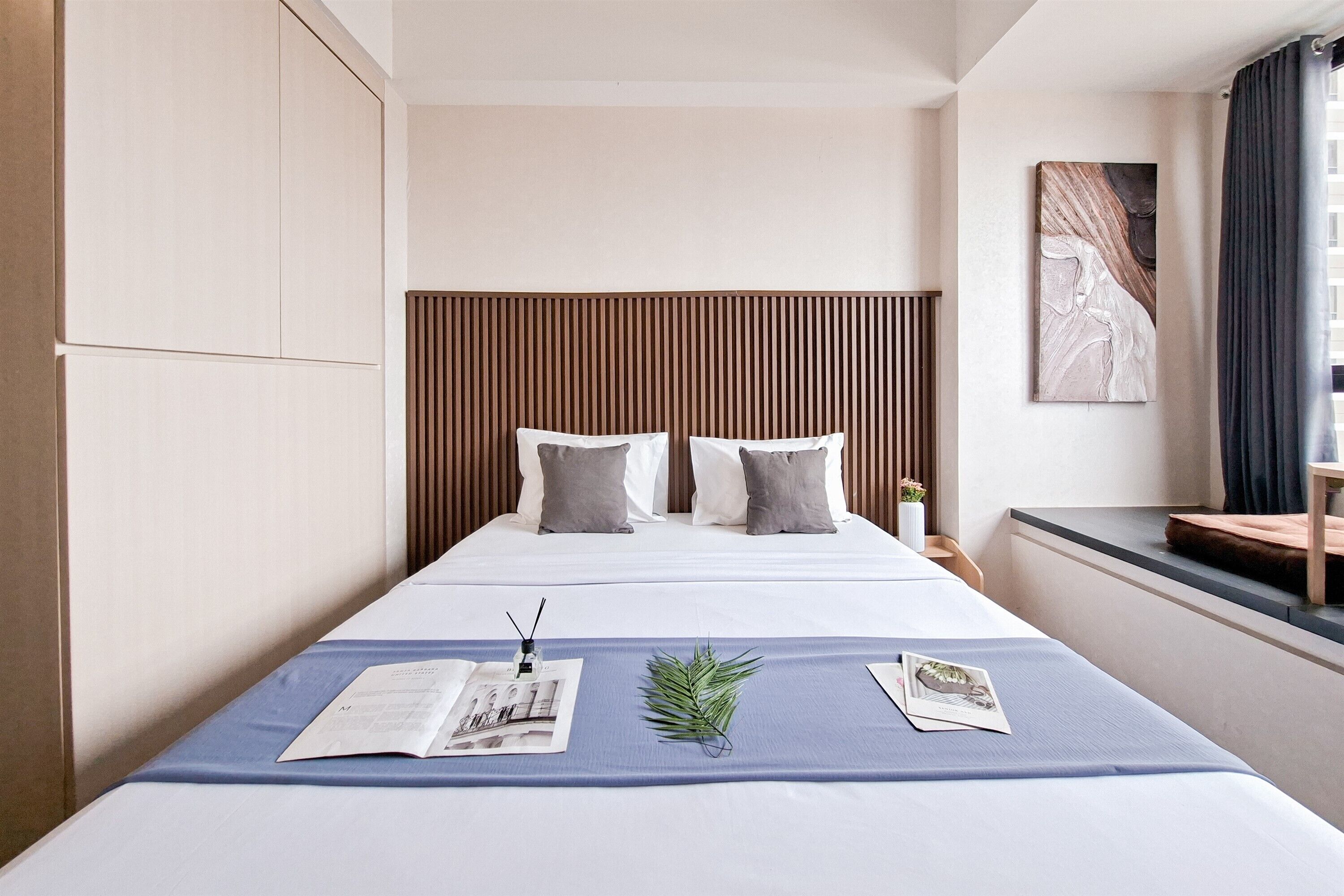 A neatly made bed with a blue bedspread, two pillows, and a small plant. A nightstand with a lamp and a book is beside the bed.