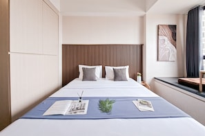 A neatly made bed with a blue bedspread, two pillows, and a small plant. A nightstand with a lamp and a book is beside the bed. - Modern Look Studio At Apartment Collins Boulevard (Tangerang)