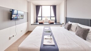 Een moderne hotelkamer met een groot bed, een flatscreen televisie aan de muur, een raam met gordijnen en een klein tafeltje bij het raam.