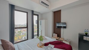 A hotel room with a large window, a bed with white linens, a red throw blanket, a small plant, and a desk with a computer and a water bottle. - Simple Studio At The Trans Icon Surabaya Apartment (Surabaya)