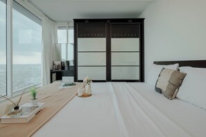 A modern hotel room with a large bed, a bedside table with a book and a vase of flowers, a wardrobe with a view, and a large window overlooking the city. - Spacious 2Br Near Mall At Grand Kamala Lagoon Apartment (Bekasi)