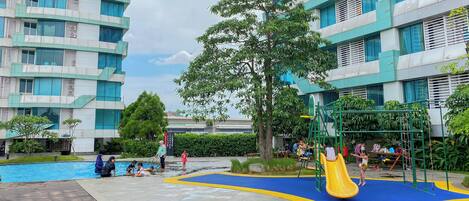 Un espace de jeux pour enfants avec un toboggan et des structures d’escalade, entouré d’une piscine et de verdure.