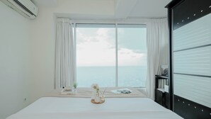 Ein Hotelzimmer mit einem großen Bett, einem Fenster mit Vorhängen, Meerblick, einem kleinen Tisch mit einer Vase und einem schwarzen Kleiderschrank.