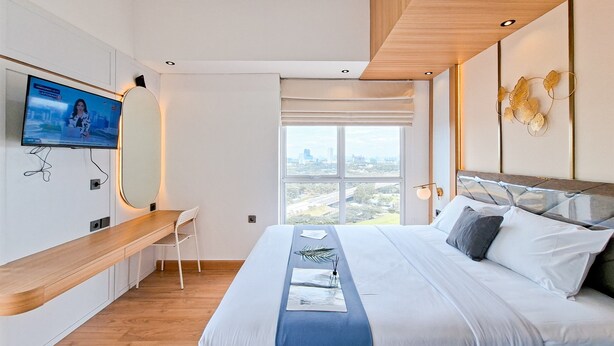 Une chambre d’hôtel moderne dotée d’un grand lit, d’un téléviseur fixé au mur, d’un bureau en bois et d’une grande fenêtre donnant sur la ville.