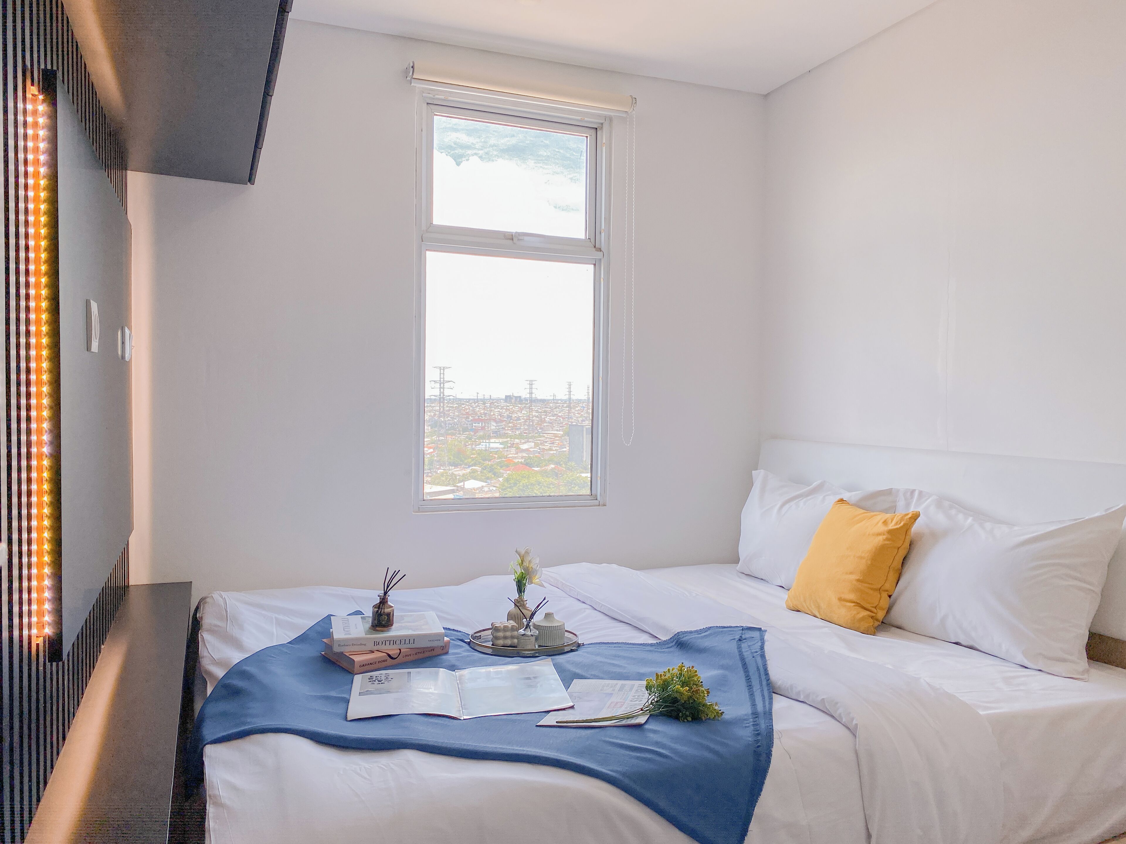Een slaapkamer met een bed, een raam met uitzicht op de stad, en een nachtkastje met boeken en een vaas.