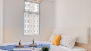 Una cama bien tendida con una manta azul, almohadas blancas y una almohada decorativa amarilla. Una mesita de noche junto a la cama sostiene un jarrón con flores, una bandeja con velas y algunos libros.