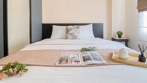 A neatly made bed with a magazine, a small plant, and a tray with a hat and reed diffuser in a modern hotel room. - Standard 2Br At Podomoro Golf View Apartment (Gunung Putri)