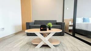 Habitación de hotel moderna con una mesa de centro de madera, un sofá gris, una planta pequeña en maceta y una cama con ropa de cama blanca.