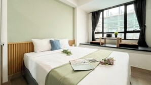 Una cama bien hecha con almohadas, un plaid verde y una revista sobre la cama. Se ve una ventana con vistas a un edificio.