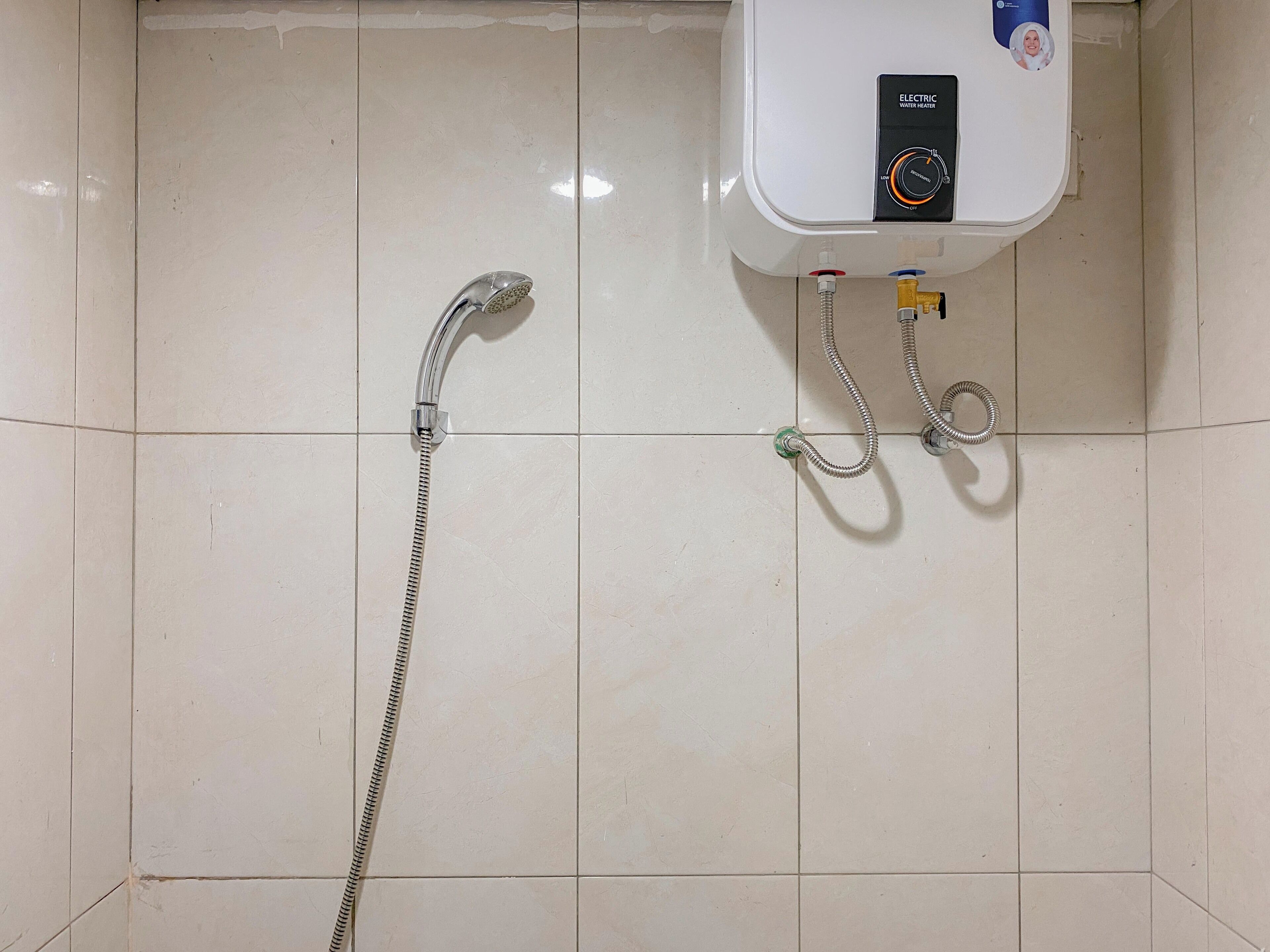 Un baño con pared de azulejos blancos, ducha de mano y calentador de agua eléctrico.
