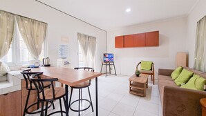 A modern living room with a wooden dining table, bar stools, a sofa with green cushions, and a television on a stand. - Modern 4Br Apartment At Guest House Vanya Park (Pagedangan)
