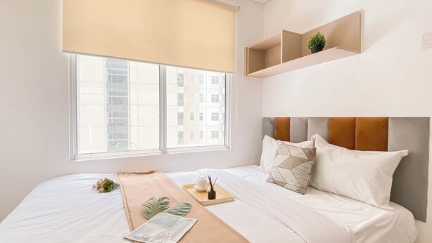 Une chambre moderne avec un grand lit, une fenêtre avec des stores, une table de chevet agrémentée d’une plante et une tête de lit ornée d’un motif géométrique.