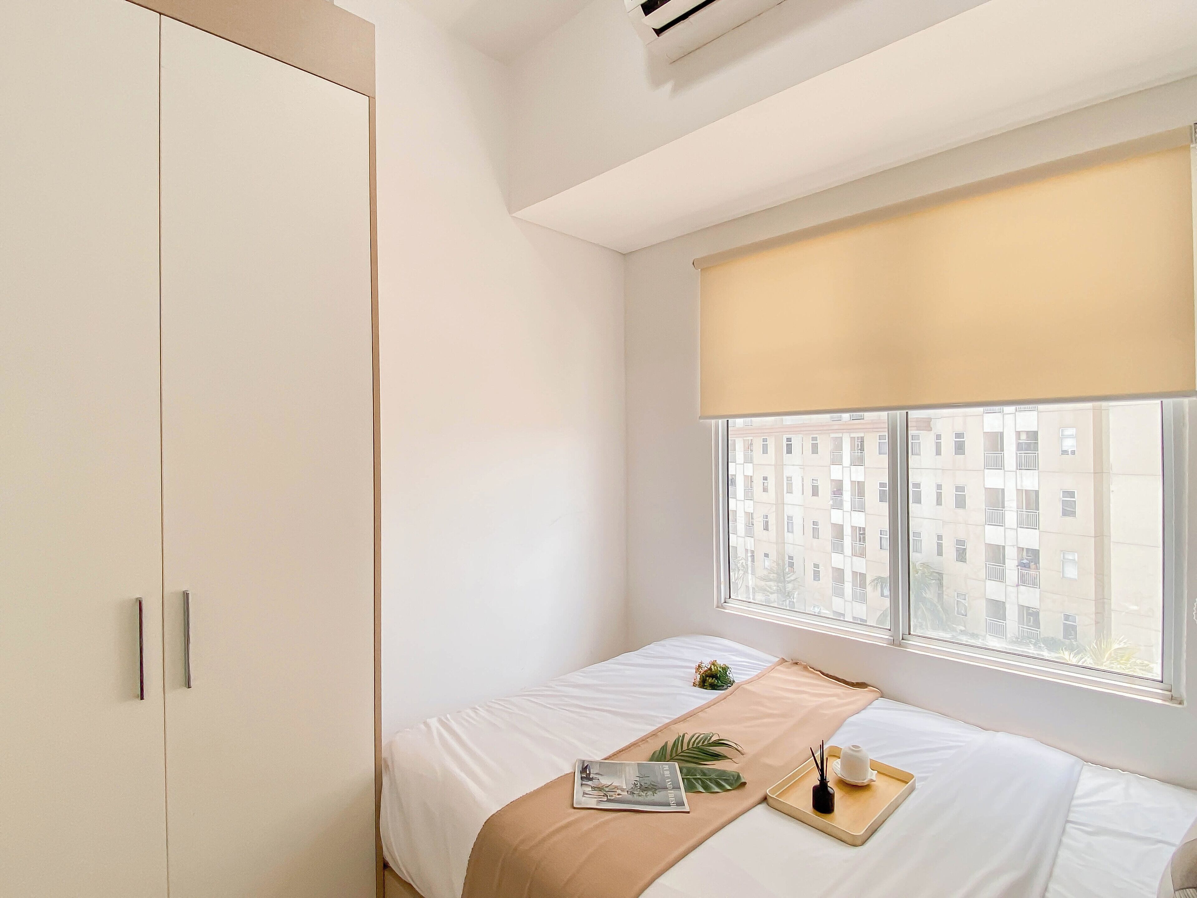 A minimalist hotel room with a bed, a window with a view of buildings, and a wardrobe.