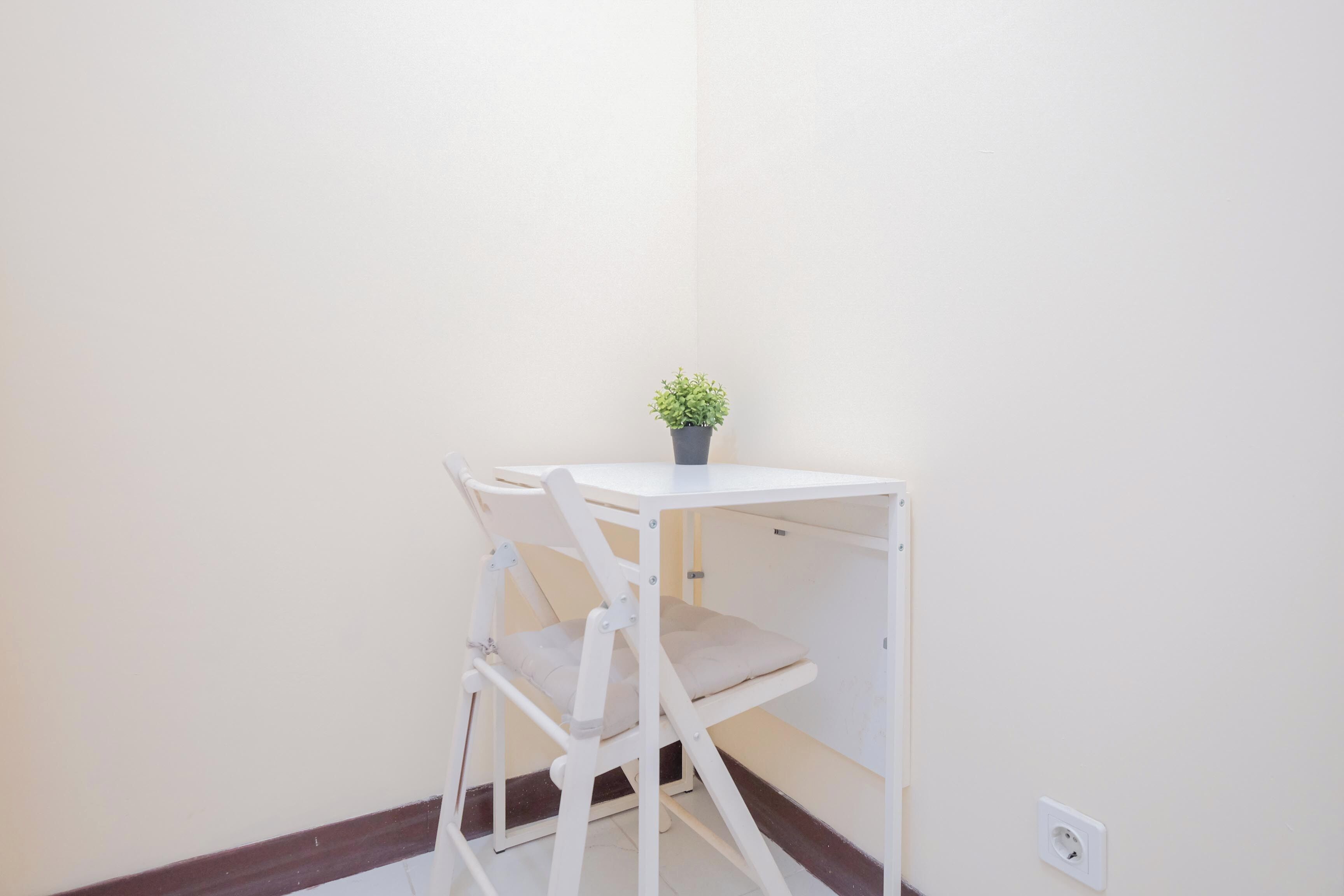 Una pequeña mesa blanca con una planta en maceta, una silla blanca y un estante blanco frente a una pared lisa.