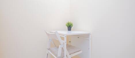 Una pequeña mesa blanca con una planta en maceta, una silla blanca y un estante blanco frente a una pared lisa.