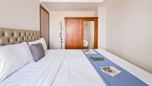 A neatly made bed with a tufted headboard, a blue bedspread with a plant, and a wooden wardrobe in the background. - Trendy 1Br Apartment At Anwa Residence (South Tangerang)