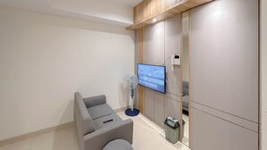 A modern room with a grey sofa, a standing fan, a television on the wall, and a small trash bin. - Cozy And Homey 2Br At Grand Sungkono Lagoon Apartment (Surabaya)