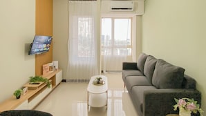 A modern living room with a flat-screen TV mounted on the wall, a grey sofa, a small white table, and a window with sheer curtains. - Basic Style And Homey 2Br At Apartment Nifarro Park (Jakarta)