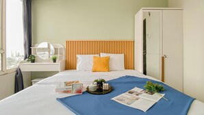 A hotel room with a bed, a bedside table with a lamp, a window with curtains, and a wardrobe. - Basic Style And Homey 2Br At Apartment Nifarro Park (Jakarta)