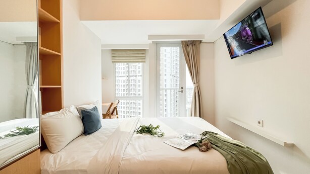 Ein Schlafzimmer mit einem Bett, einem an der Wand befestigten Fernseher, einem Fenster mit Vorhängen und Blick auf Gebäude.