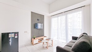 Ein modernes Wohnzimmer mit einem grauen Sofa, einem kleinen runden Tisch, einem schwarzen Kühlschrank, einer Holzkabine und einem großen Fenster mit durchscheinenden Vorhängen.