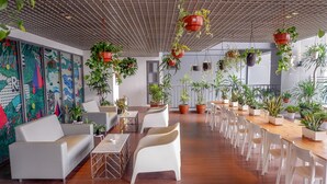 Un espacio interior amplio con una larga mesa de madera y sillas blancas, rodeado de plantas en macetas y un mural colorido en la pared.