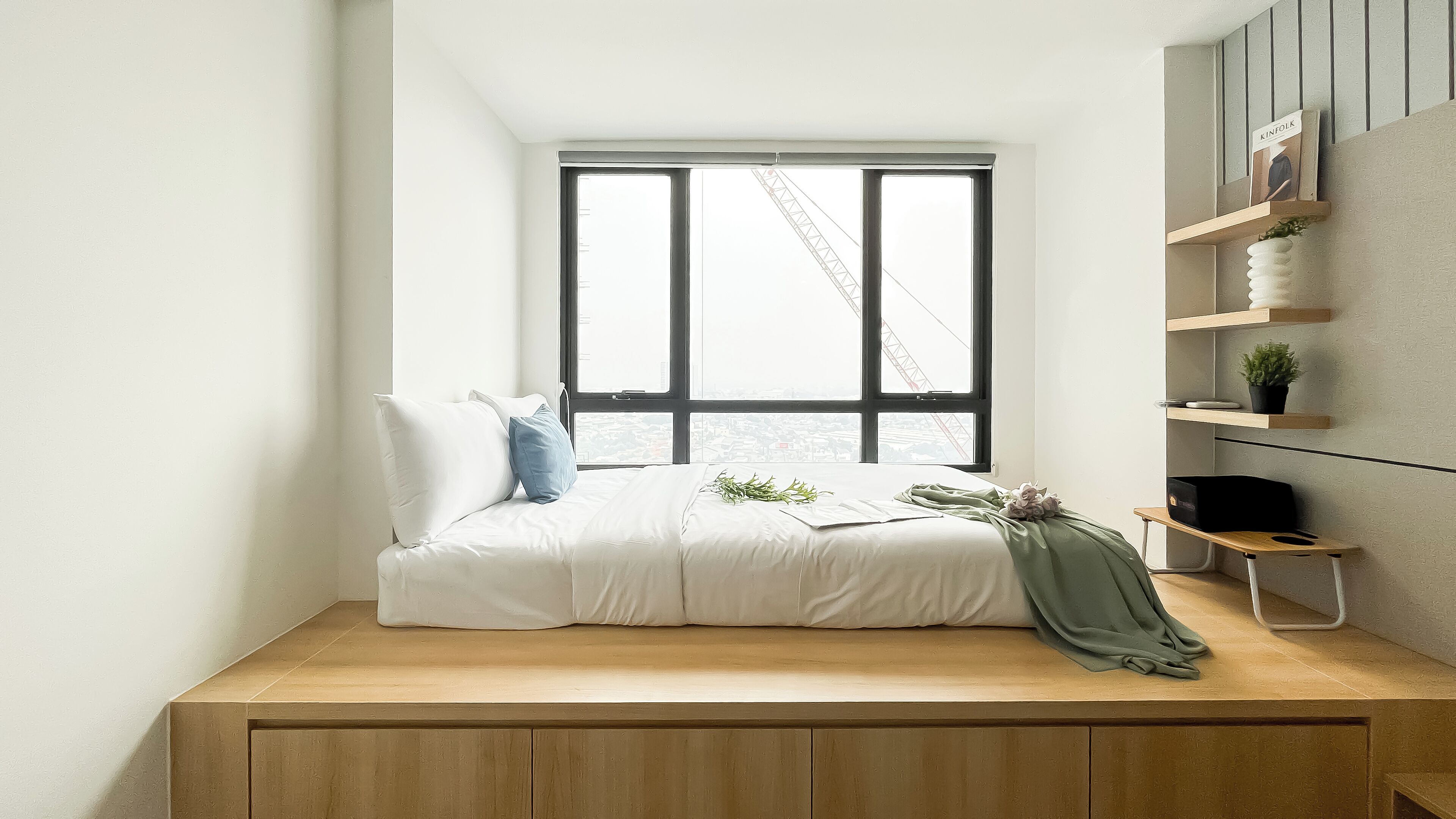 A bedroom with a bed, a window with a view of a crane, and a shelf with plants.