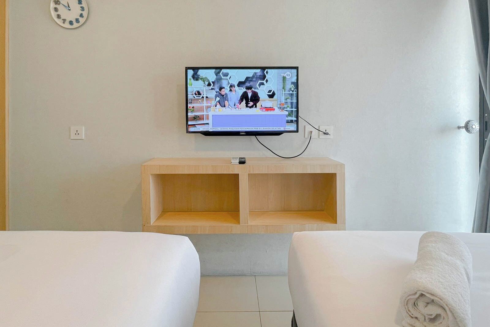 A hotel room with a television mounted on the wall, a wooden shelf below it, and two beds with white linens.