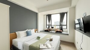 Una habitación de hotel con una cama, un ventanal con cortinas, un televisor montado en la pared y una mesita con un florero y un libro.