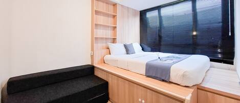 A compact bedroom with a bed, built-in shelving, a window with blinds, and a black sofa.