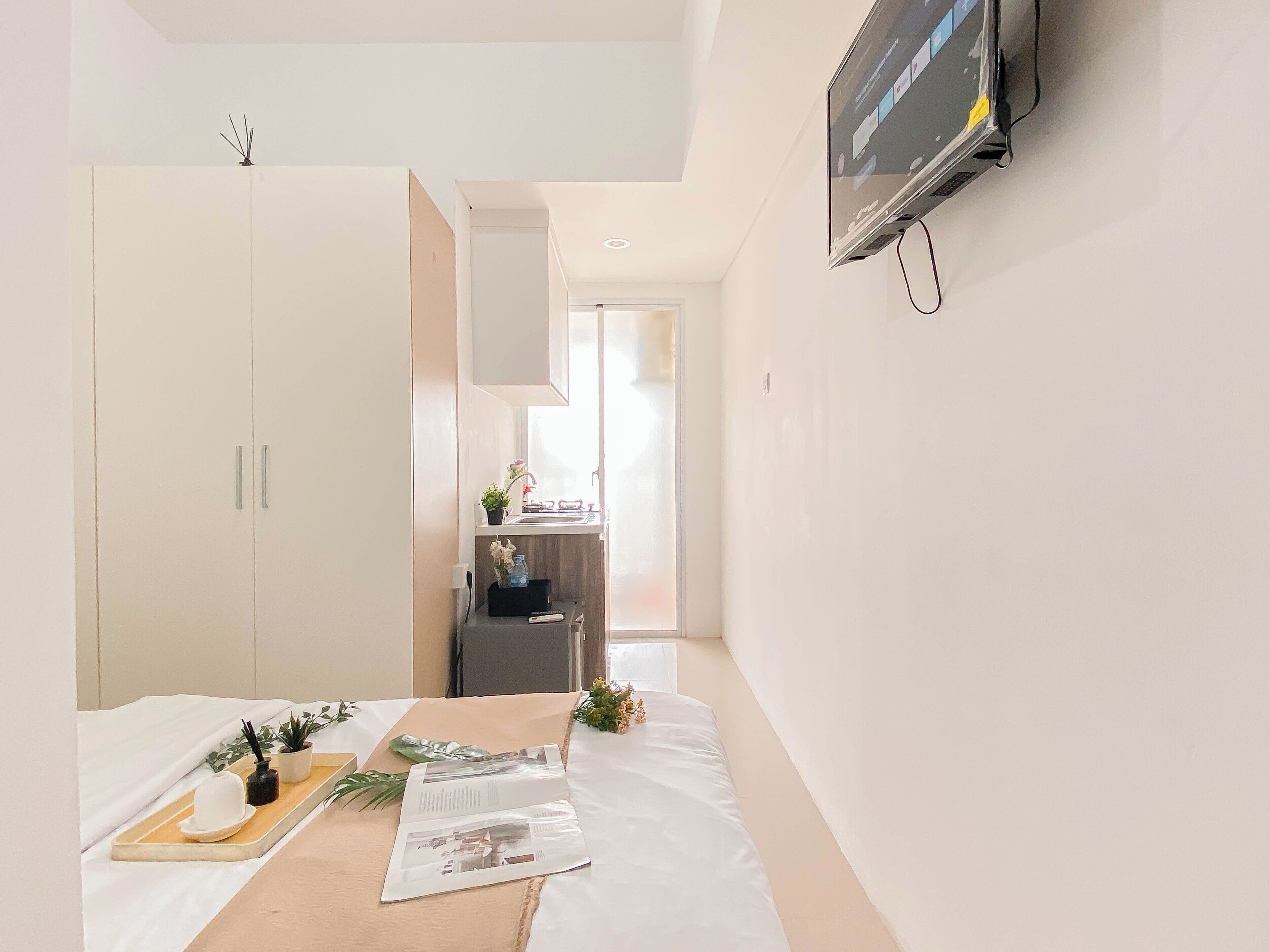 A hotel room with a bed, a small table with magazines, a TV mounted on the wall, and a kitchenette area.