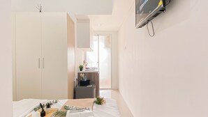 A hotel room with a bed, a small table with magazines, a TV mounted on the wall, and a kitchenette area. - Basic Studio Apartment At Vittoria Residence (Jakarta)