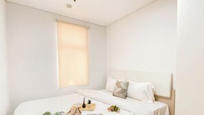 A neatly made bed with white linens, a bedside table with a tray, and a window with blinds. - Ivory Nest 1Br At Vittoria Residence Apartment (Jakarta)