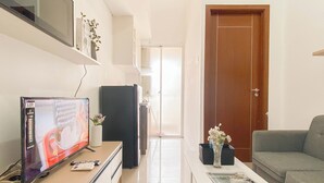 A modern living room with a flat-screen TV, a refrigerator, a coffee table with a magazine, a grey sofa, and a door leading to another room. - Chic 1Br Apartment At Vittoria Residence (Jakarta)