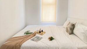 A bedroom with a bed, a window with blinds, a bedside table with a plant, a book, and a decorative pillow. - Chic 1Br Apartment At Vittoria Residence (Jakarta)